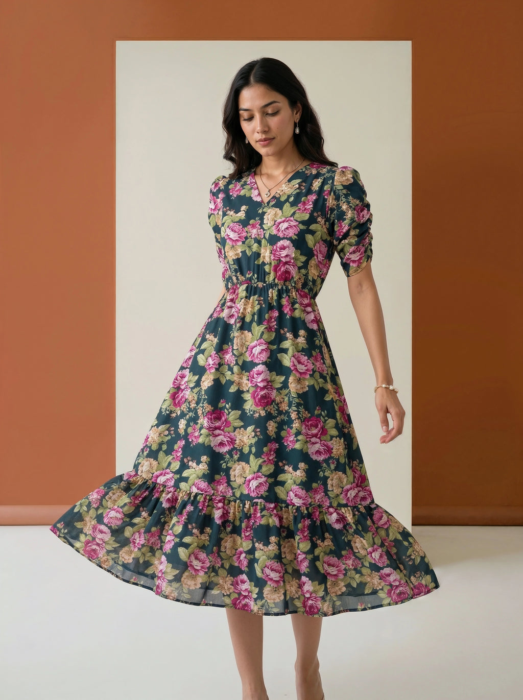 A woman modeling a dark green midi dress with a pink and yellow floral print, standing in front of a minimalist studio backdrop with a warm terracotta wall and cream panel.