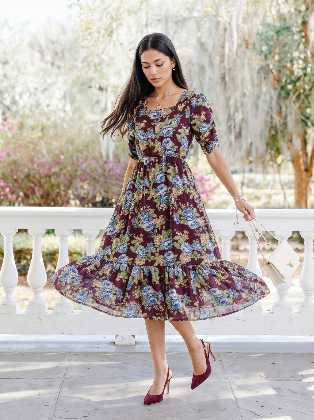 A woman wearing a short-sleeve, tiered midi dress with a dark purple and blue floral print, standing outdoors in front of a white railing.