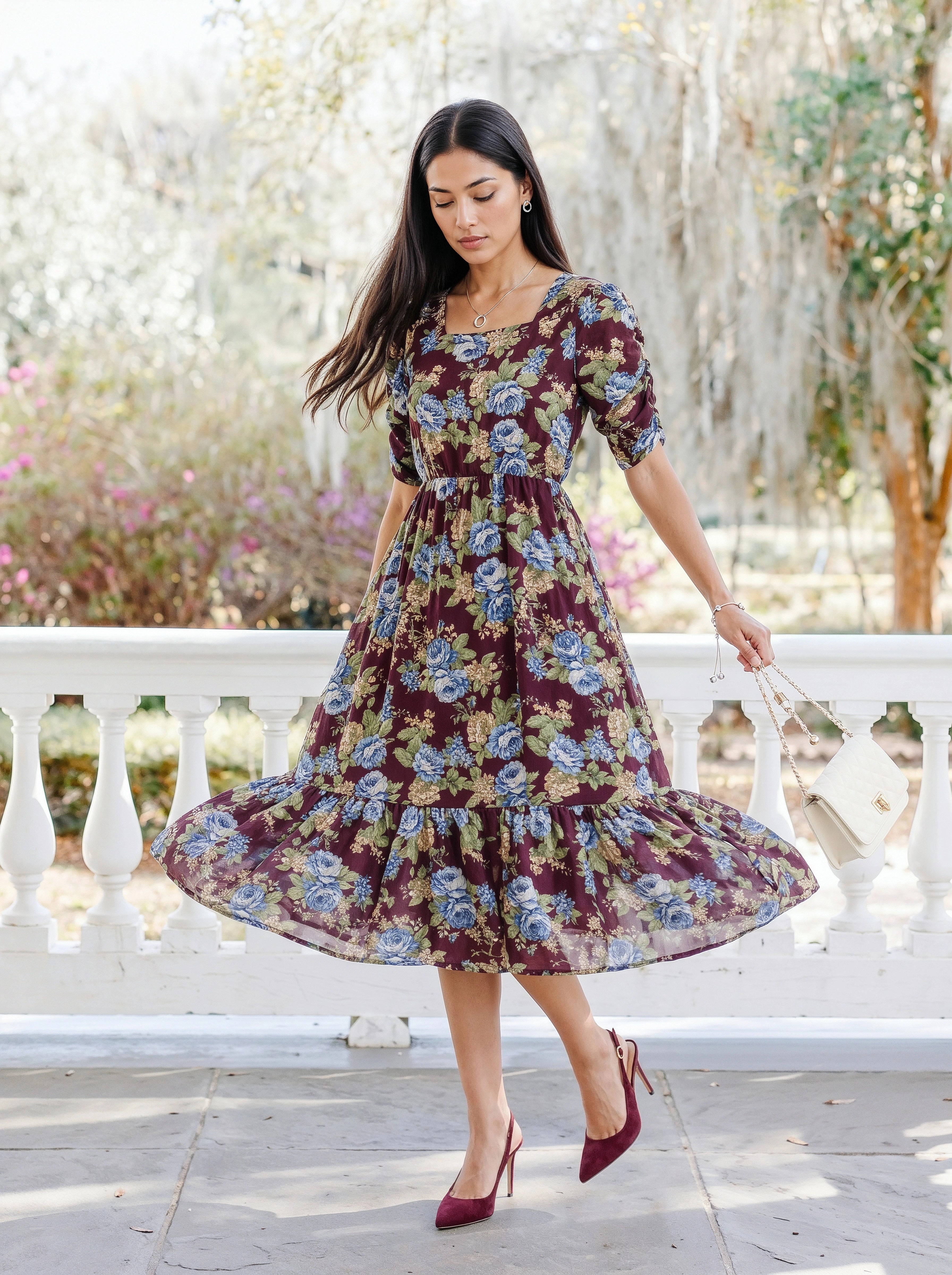 A woman wearing a short-sleeve, tiered midi dress with a dark purple and blue floral print, standing outdoors in front of a white railing.
