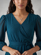Woman wearing a teal dress with long sleeves and a necklace on a neutral background