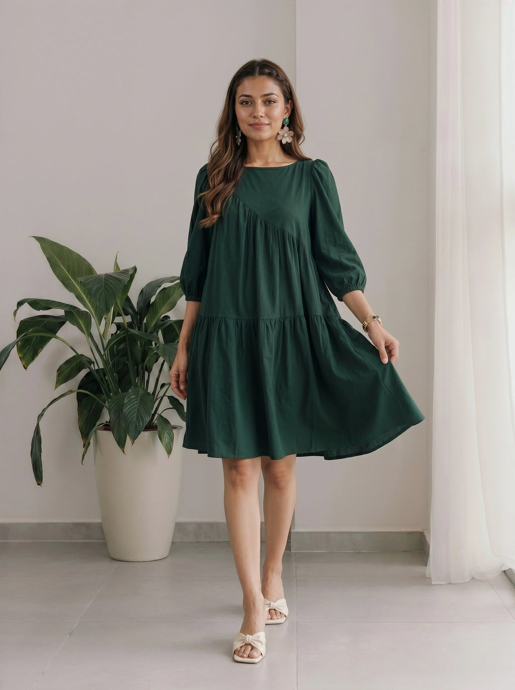 Woman wearing a green dress standing indoors with a plant and curtain in the background