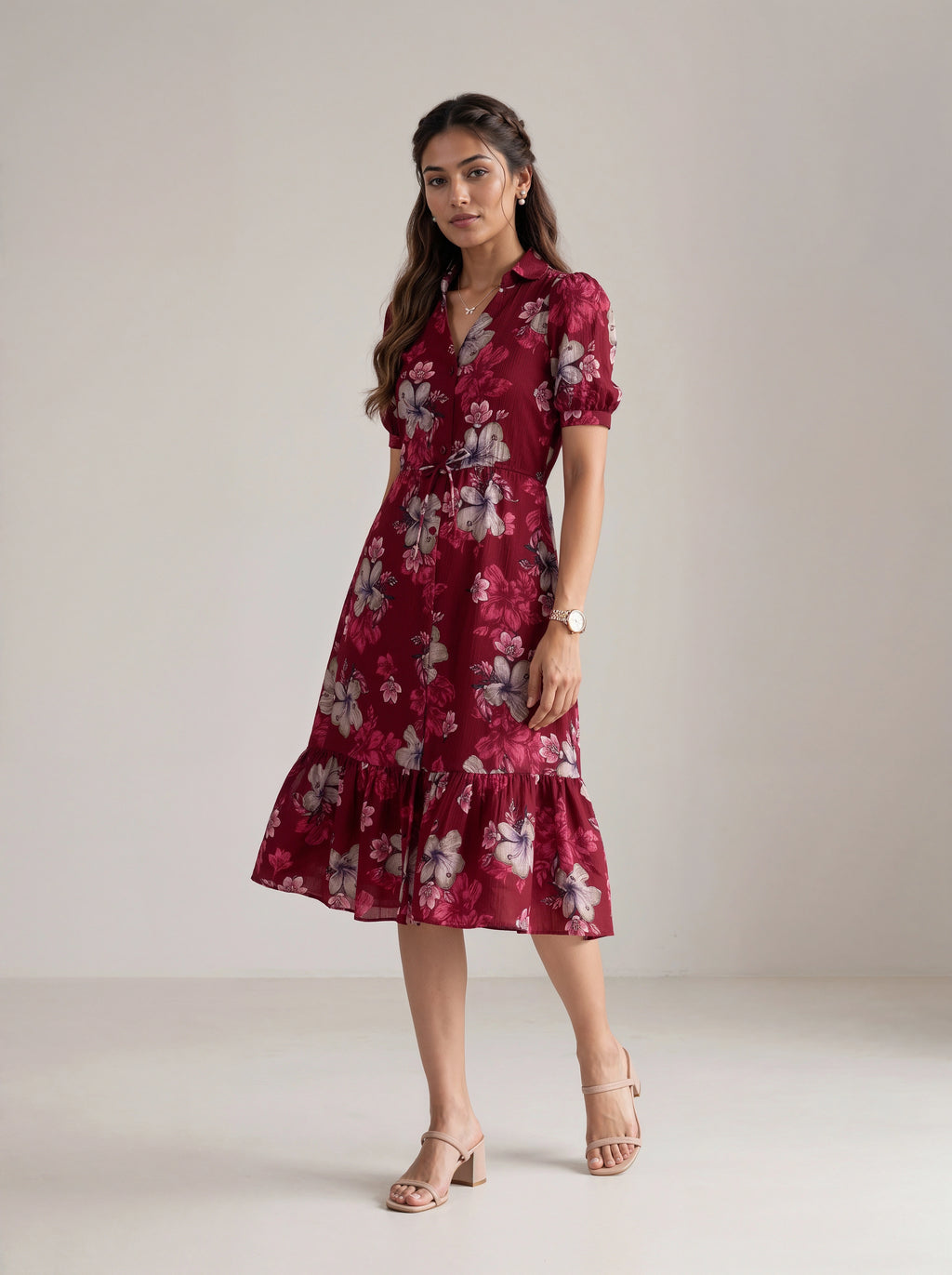 A woman modeling a short-sleeve midi dress with a floral print in deep red, burgundy, and white, paired with beige heeled sandals against a neutral studio backdrop.