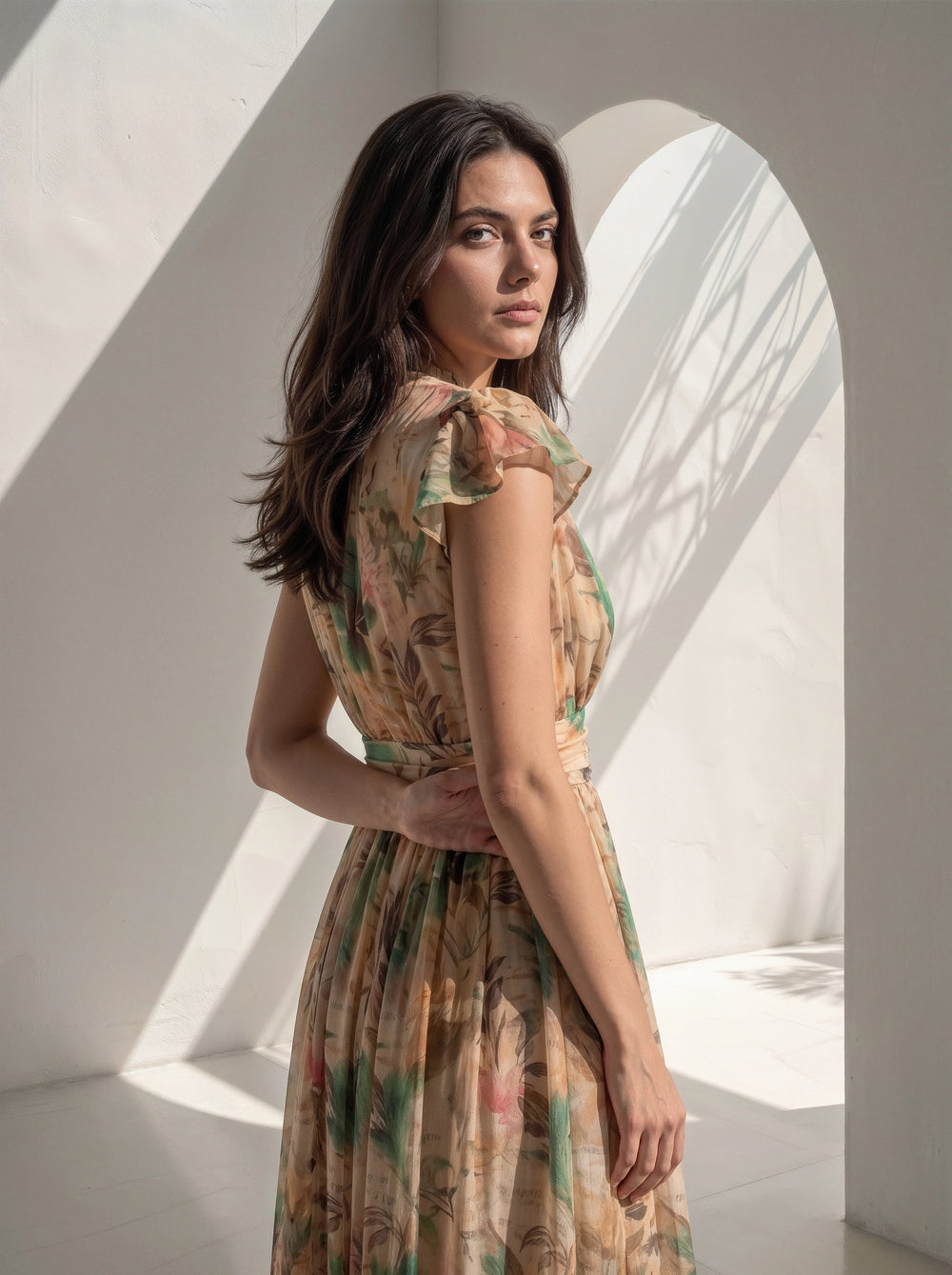 A woman wearing a sleeveless, knee-length dress with a botanical floral print in beige, green, and pink, standing in a bright white architectural setting with dramatic shadows.