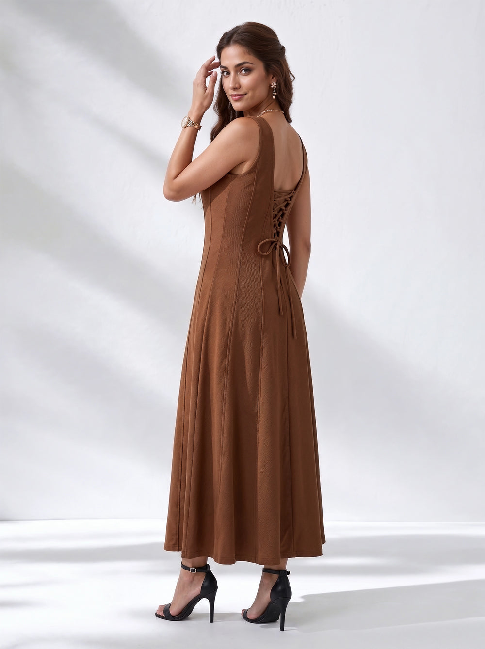 A woman modeling a sleeveless brown maxi dress from the back, featuring a lace-up open back and tie detail, paired with black high-heeled sandals against a white studio backdrop with soft shadows.