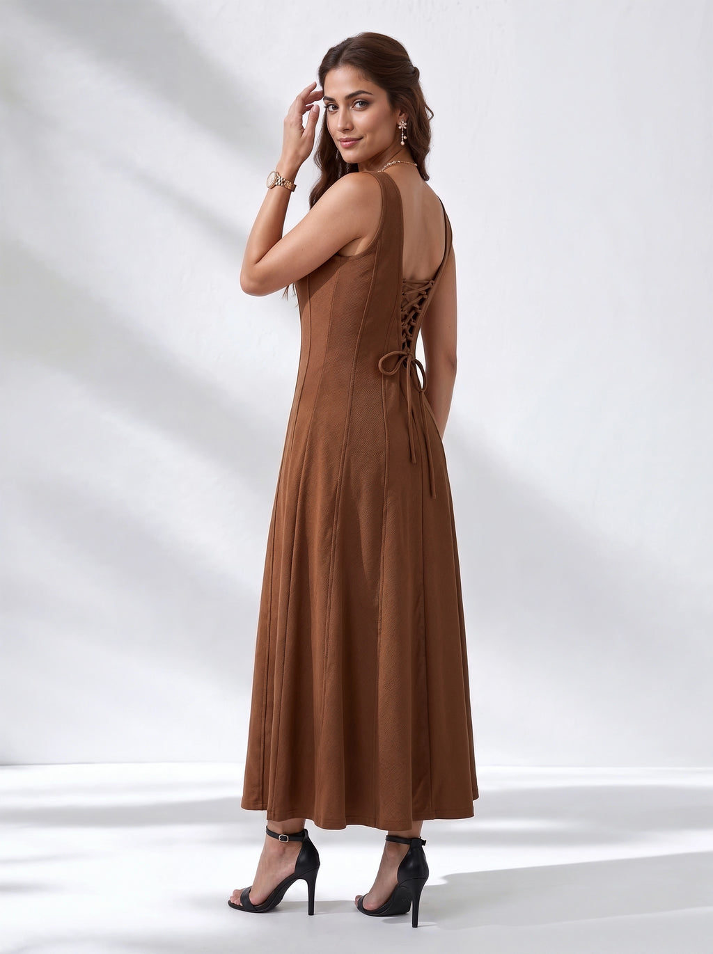 A woman modeling a sleeveless brown maxi dress from the back, featuring a lace-up open back and tie detail, paired with black high-heeled sandals against a white studio backdrop with soft shadows.
