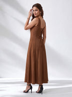 A woman modeling a sleeveless brown maxi dress from the back, featuring a lace-up open back and tie detail, paired with black high-heeled sandals against a white studio backdrop with soft shadows.