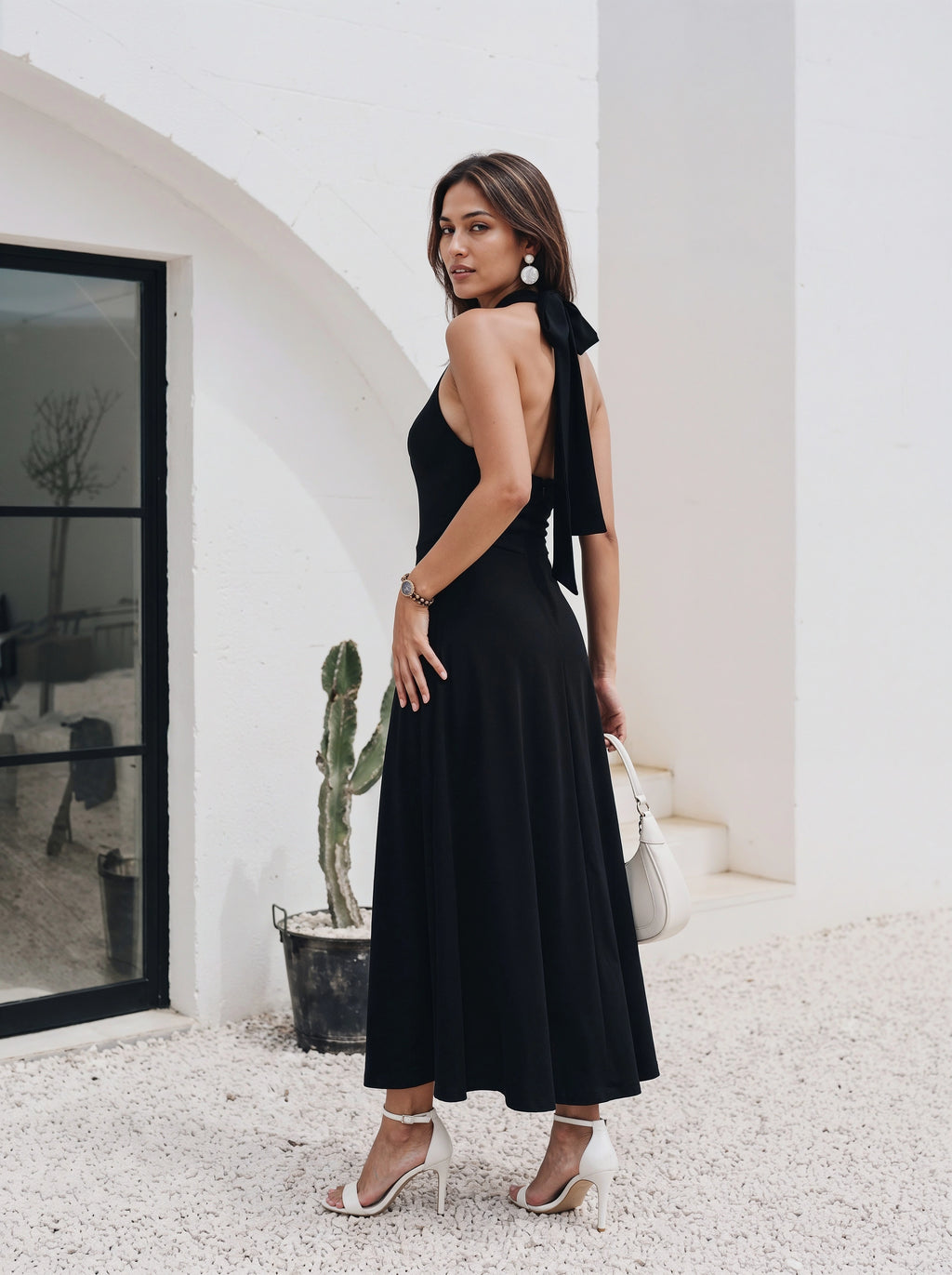 A woman models a black halter-neck maxi dress with a dramatic side bow detail and an open back, paired with white high-heeled sandals and a white handbag, set against a white stucco building background.