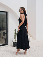 A woman models a black halter-neck maxi dress with a dramatic side bow detail and an open back, paired with white high-heeled sandals and a white handbag, set against a white stucco building background.
