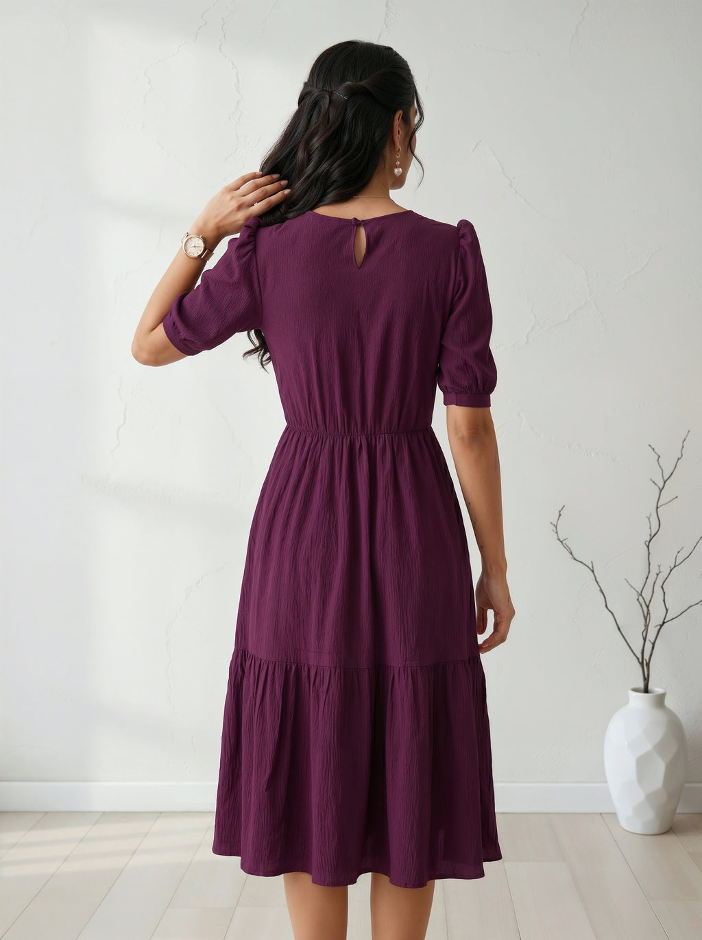 Woman wearing a purple dress standing in a minimalistic room with a white vase and plant.