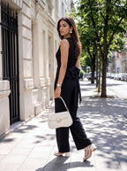 A woman walks on a sunny city sidewalk wearing a black sleeveless wide-leg jumpsuit with a large side bow, paired with a white handbag and white heeled sandals.