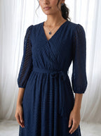 Woman wearing a navy blue dress with a textured pattern against a white curtain background
