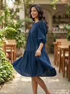 Woman in a blue dress standing in an outdoor setting with plants and tables.
