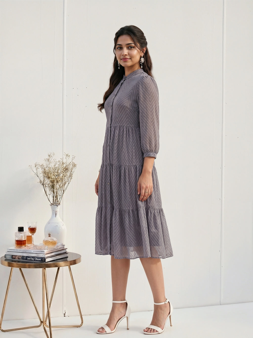 Woman in a gray dress standing next to a small table with a vase and books on a white background