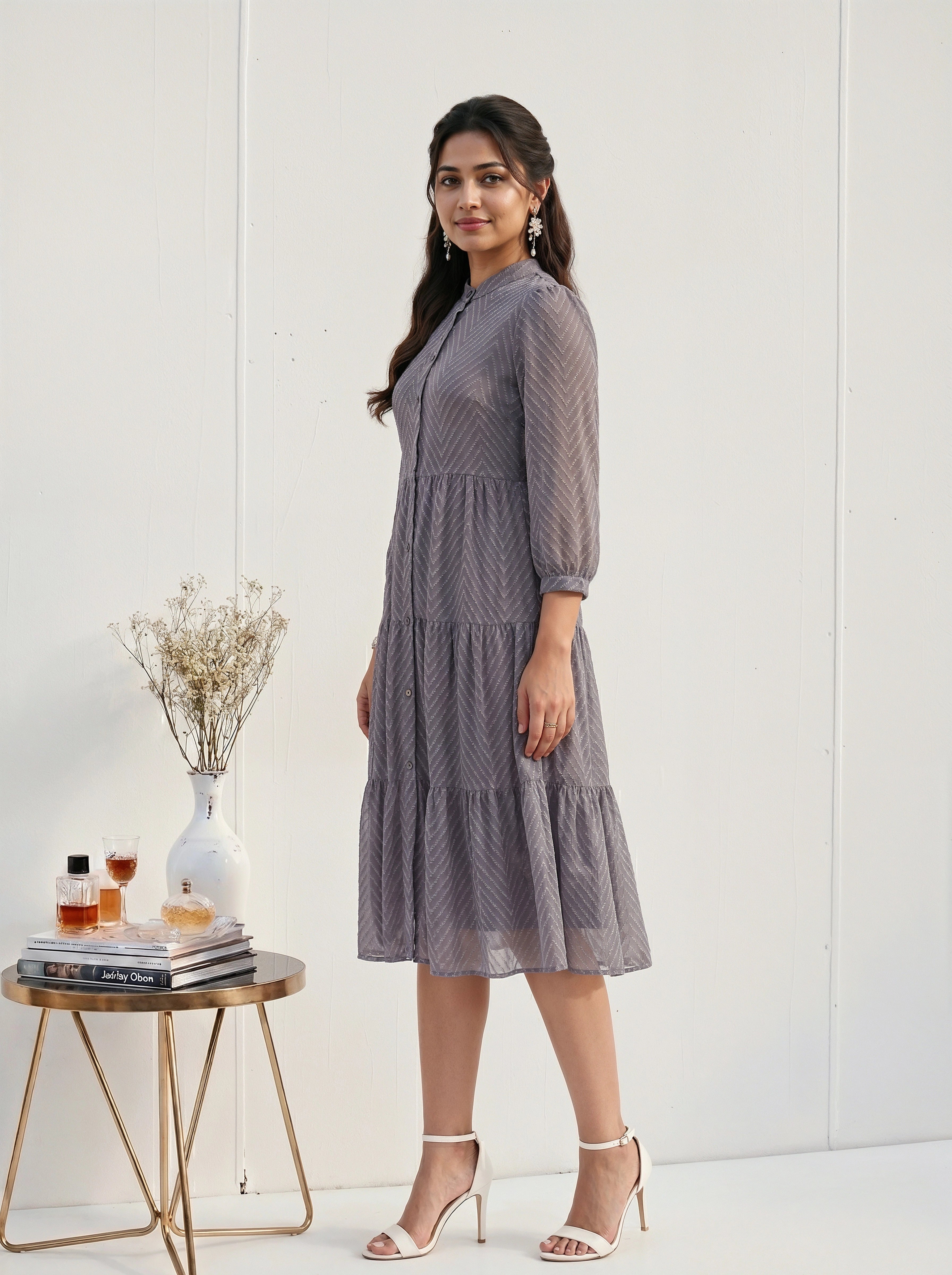 Woman in a gray dress standing next to a small table with a vase and books on a white background