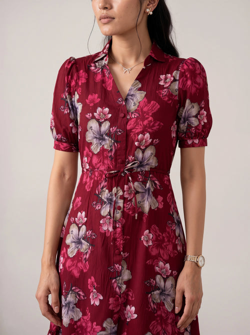 A woman wearing a short-sleeve collared dress with a red base and a large floral print in pink, purple, and gray, featuring a tie waist and button front.