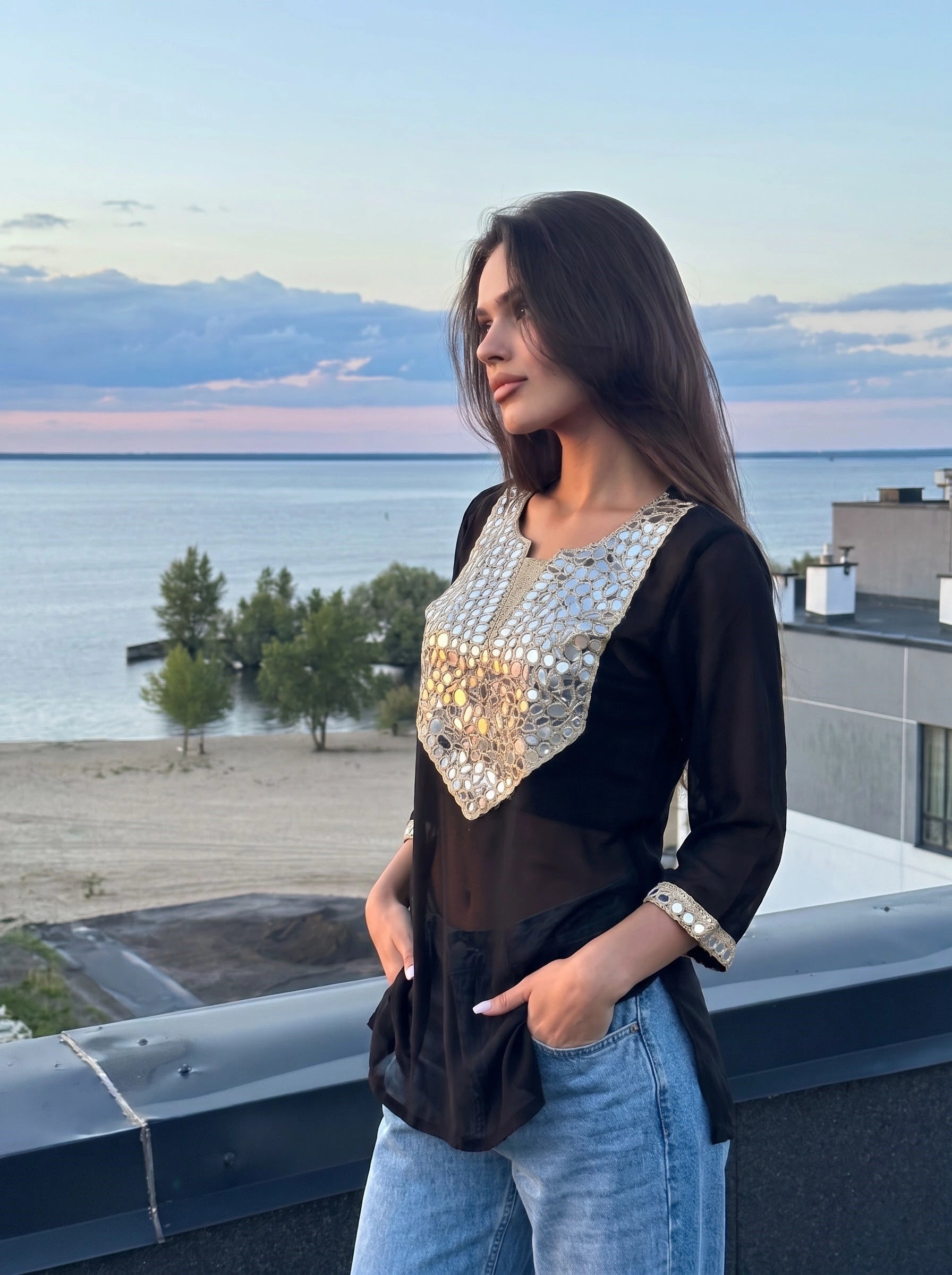 A woman wearing a black long-sleeve top with a large silver sequined yoke and light blue jeans, standing on a rooftop with a lake and beach in the background at sunset.