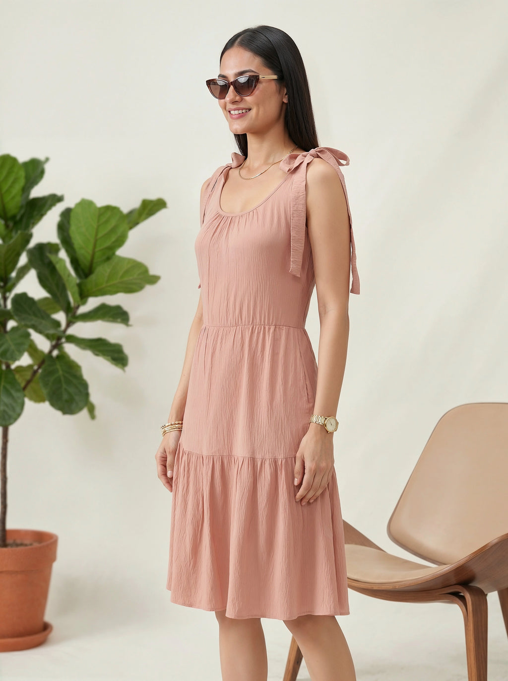 Woman wearing a pink dress standing next to a plant and chair indoors.