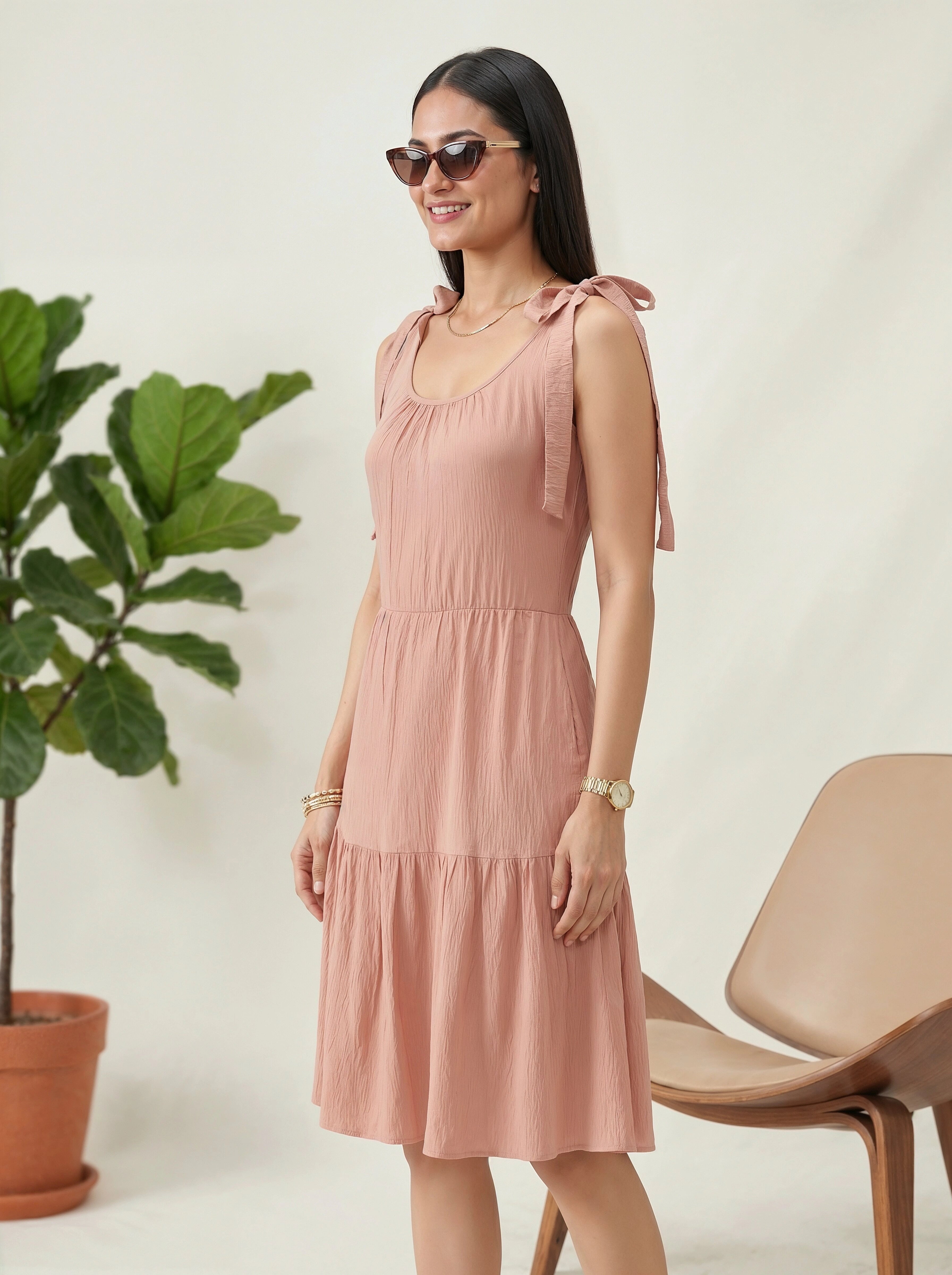 Woman wearing a pink dress standing next to a plant and chair indoors.