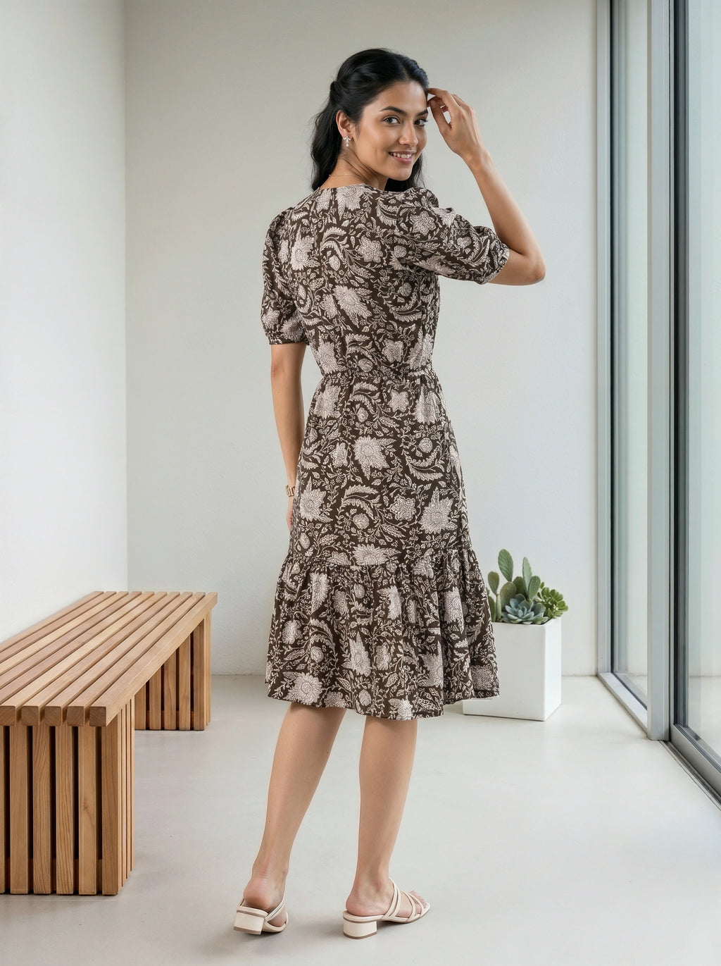A woman modeling a short-sleeve midi dress with a brown and beige paisley pattern, standing in a bright, modern room with a wooden bench and large windows.