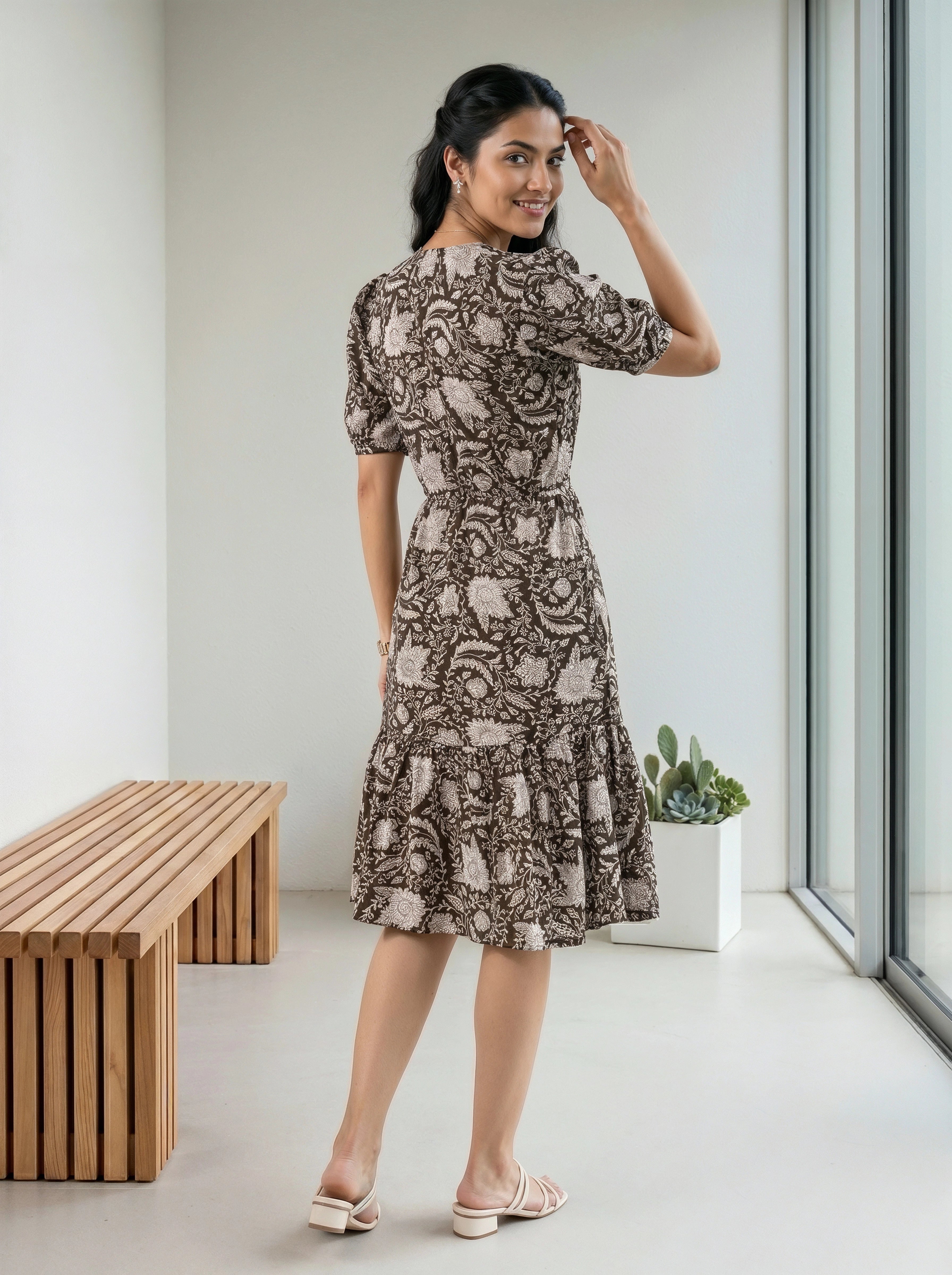 A woman modeling a short-sleeve midi dress with a brown and beige paisley pattern, standing in a bright, modern room with a wooden bench and large windows.