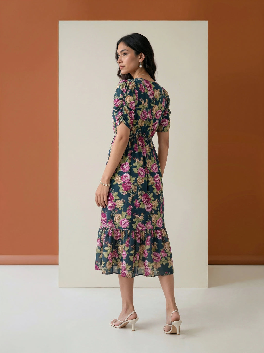 A woman modeling a dark floral midi dress with puff sleeves and a ruffled hem, standing in front of a minimalist studio backdrop with a terracotta wall and cream panel.