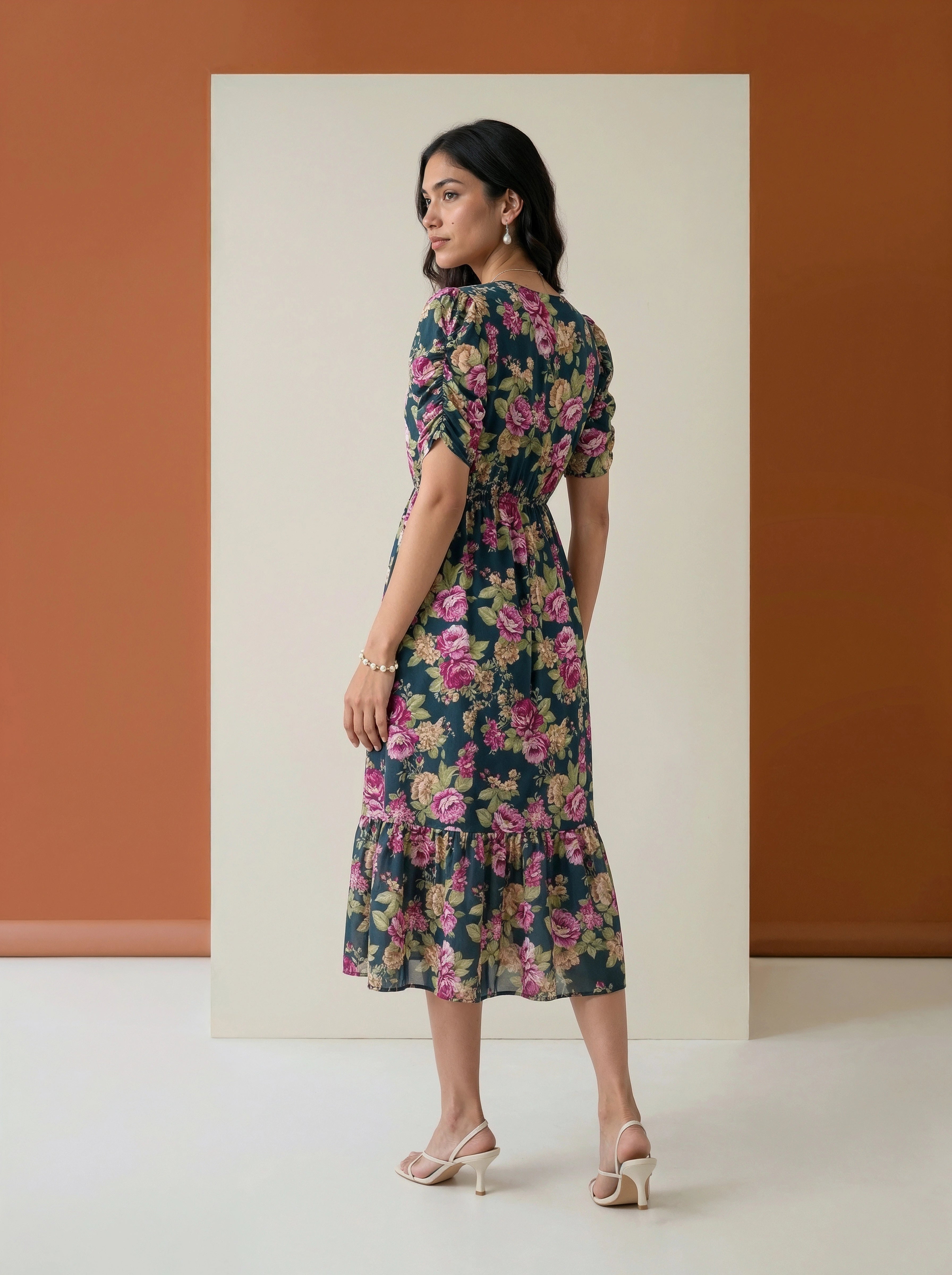 A woman modeling a dark floral midi dress with puff sleeves and a ruffled hem, standing in front of a minimalist studio backdrop with a terracotta wall and cream panel.