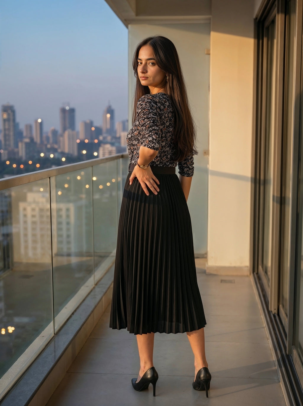 Woman in a black pleated skirt and patterned top standing on a balcony with a cityscape view.