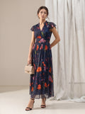 A woman models a navy blue midi dress with a vibrant orange, red, and pink floral print. The dress features short flutter sleeves, a V-neckline, and a matching fabric belt at the waist.