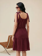 Woman wearing a burgundy dress with bow straps, standing against a beige background.
