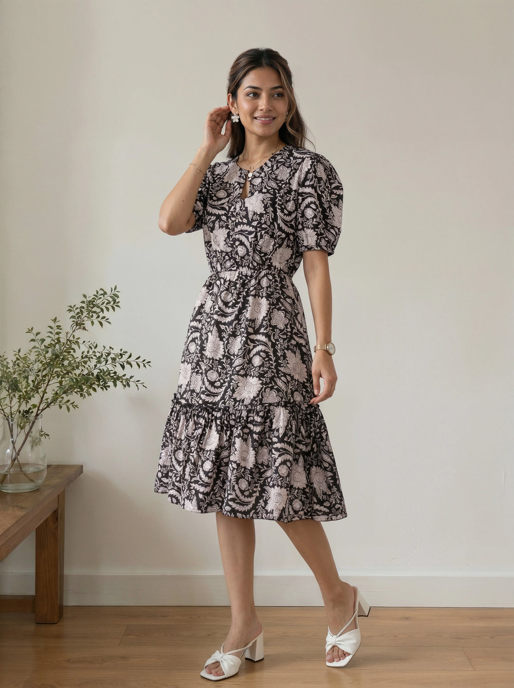A woman modeling a short puff-sleeve midi dress with a black and dusty pink paisley pattern, standing in a minimalist room with a wooden floor and a small plant on a side table.