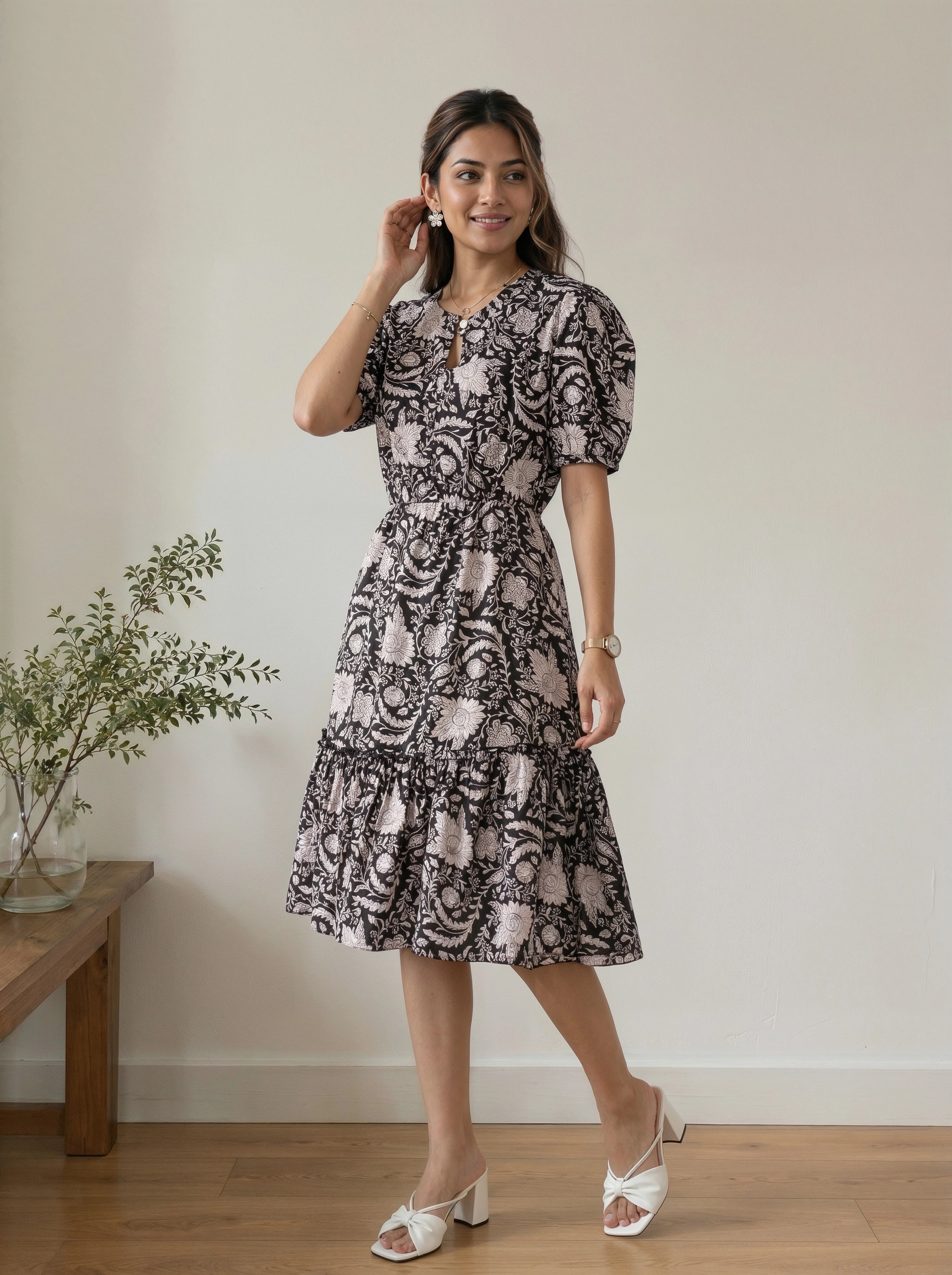 A woman modeling a short puff-sleeve midi dress with a black and dusty pink paisley pattern, standing in a minimalist room with a wooden floor and a small plant on a side table.