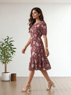 A woman modeling a short-sleeve, knee-length dress with a red and beige paisley pattern, walking in a minimalist room with a wooden floor and a potted plant.