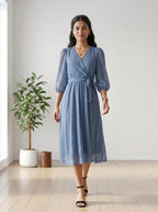 Woman wearing a blue dress standing in a room with a plant and wooden stool.