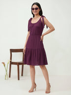 Woman wearing a purple dress standing against a white background with a wooden chair and plant.