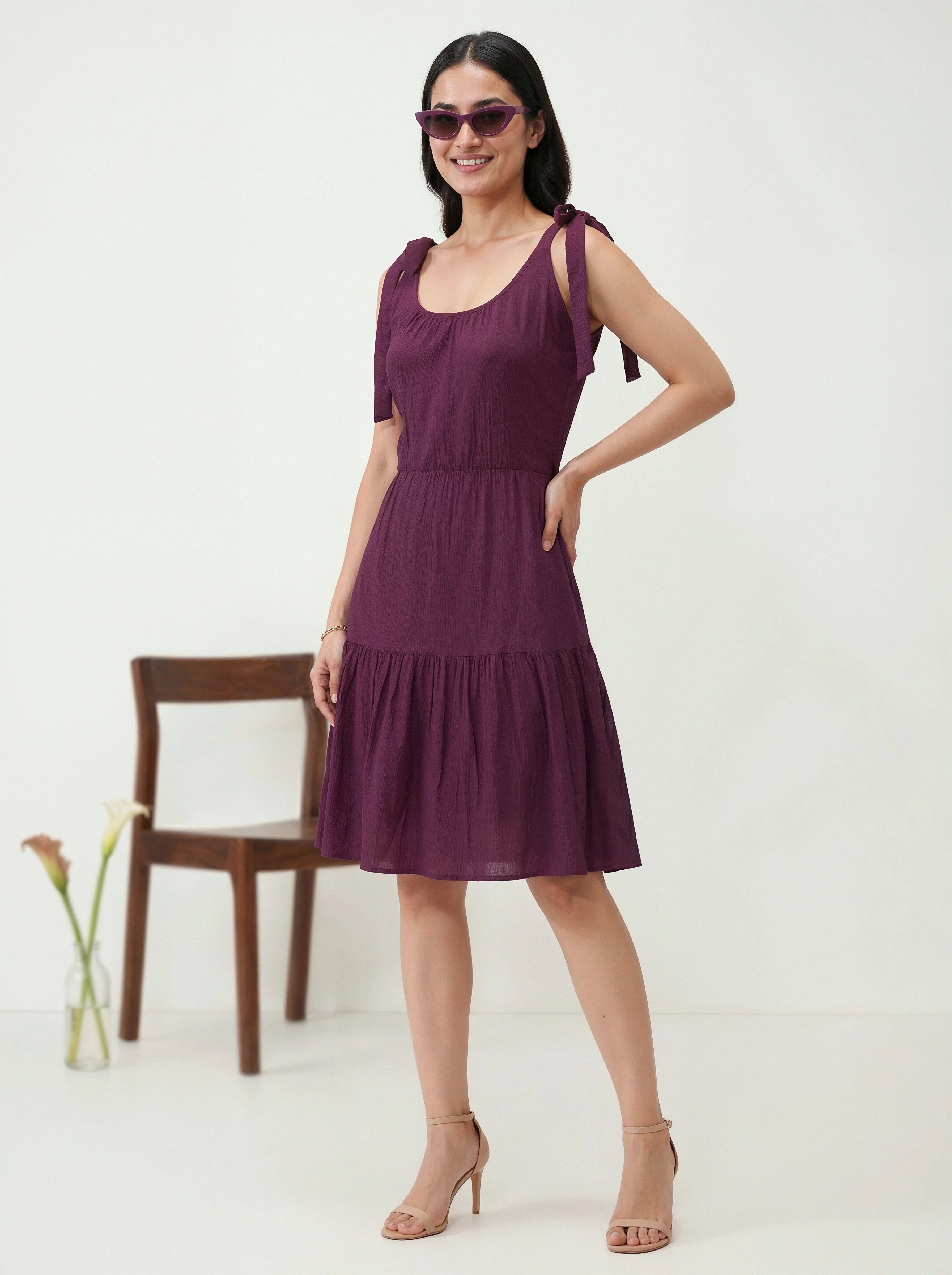 Woman wearing a purple dress standing against a white background with a wooden chair and plant.