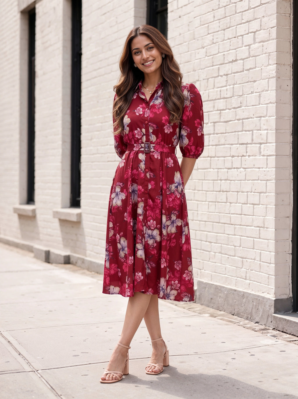 A woman stands on a sidewalk in front of a white brick building, wearing a red floral midi dress with puff sleeves and a tie waist, paired with beige heeled sandals.