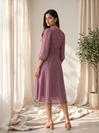 Woman in a purple dress standing in a softly lit room with curtains and a plant.
