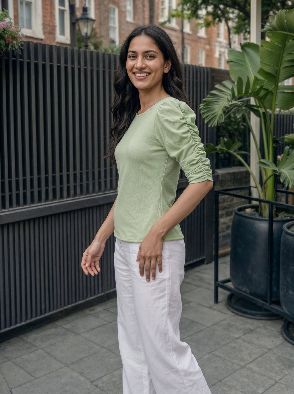Woman wearing a light green top and white pants standing outdoors with plants and a building in the background.