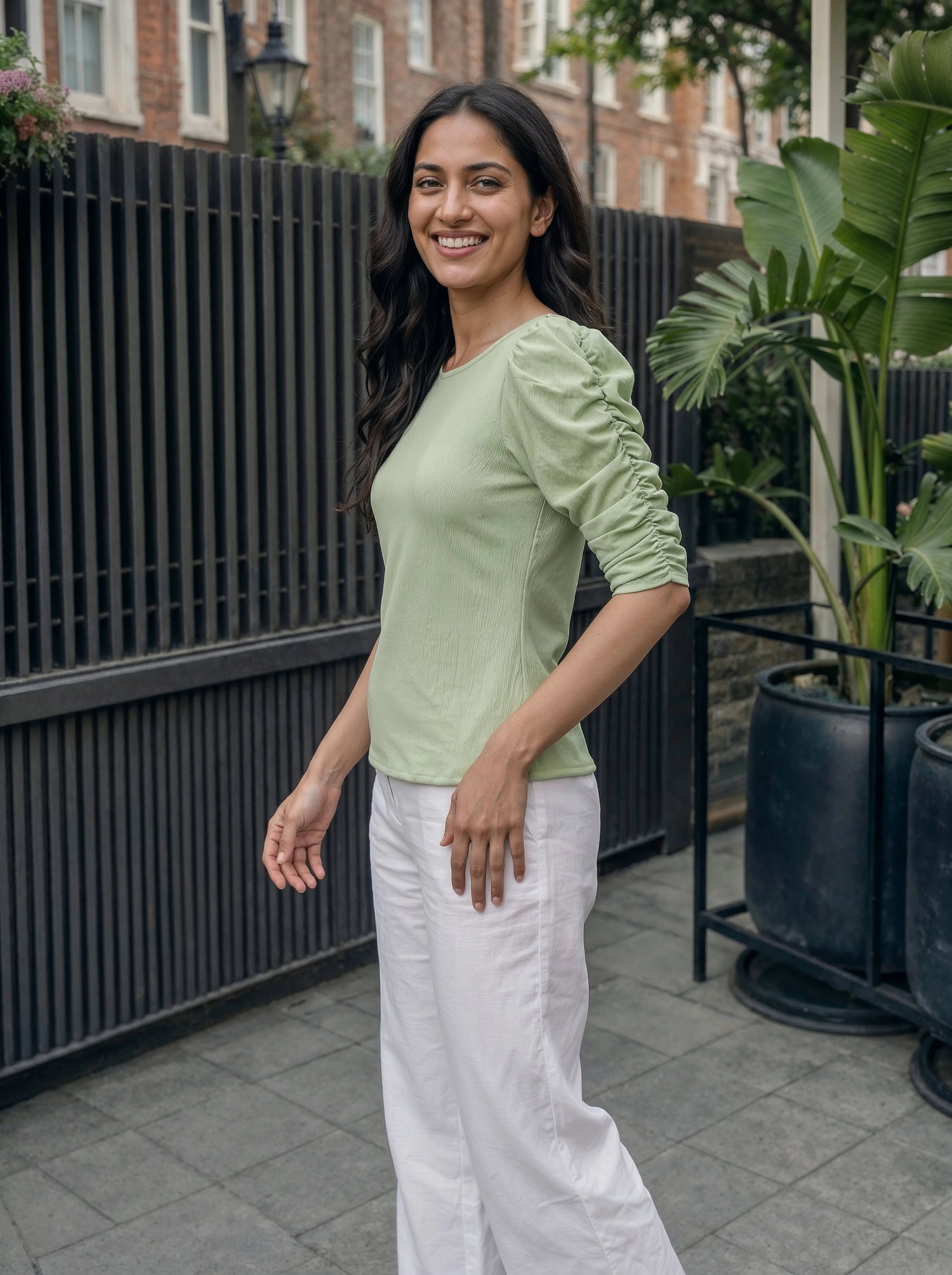 Woman wearing a light green top and white pants standing outdoors with plants and a building in the background.