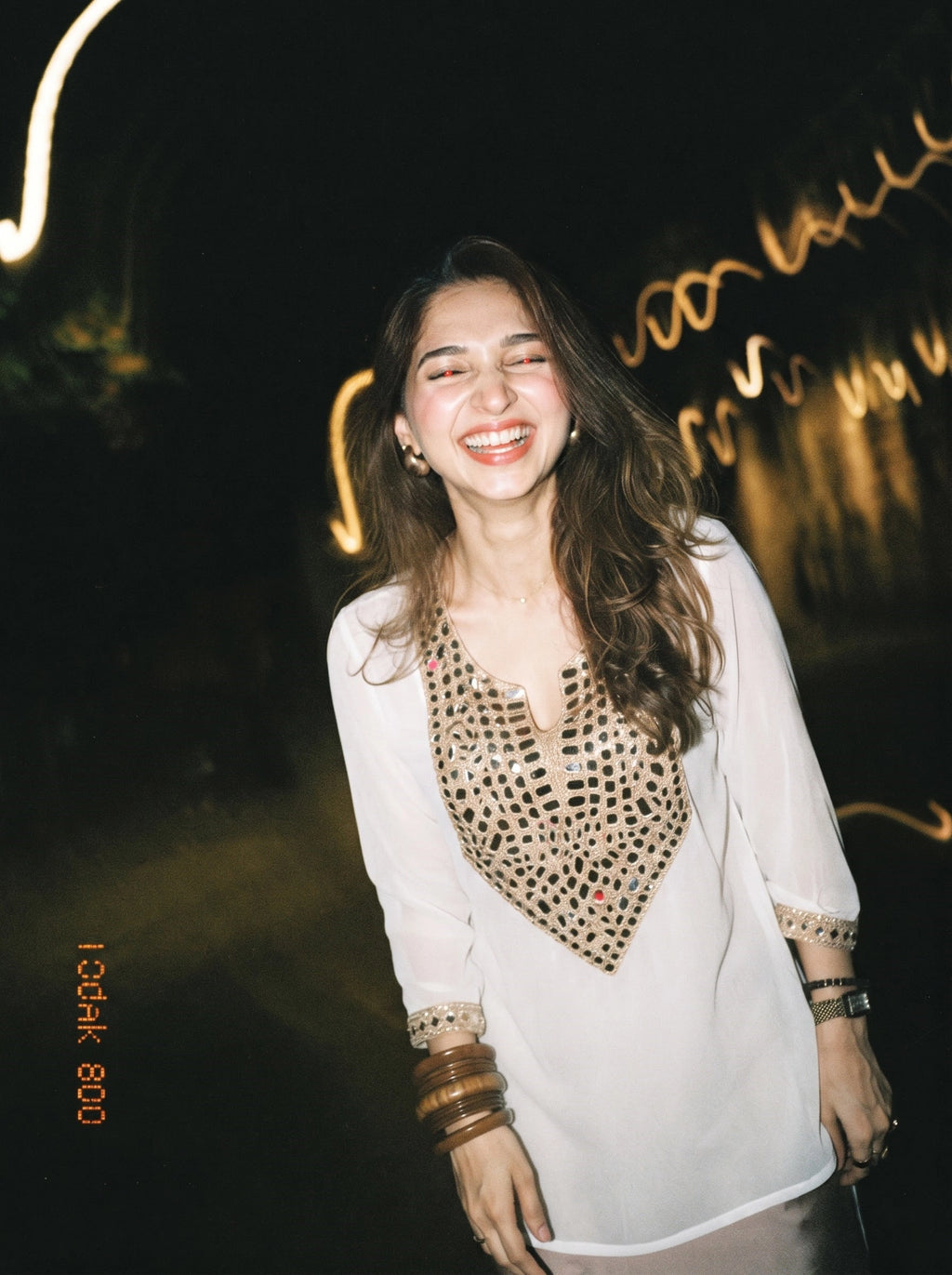 A woman smiling in a white long-sleeve tunic top with a gold geometric patterned chest panel, photographed at night with warm light trails in the background.