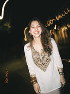A woman smiling in a white long-sleeve tunic top with a gold geometric patterned chest panel, photographed at night with warm light trails in the background.