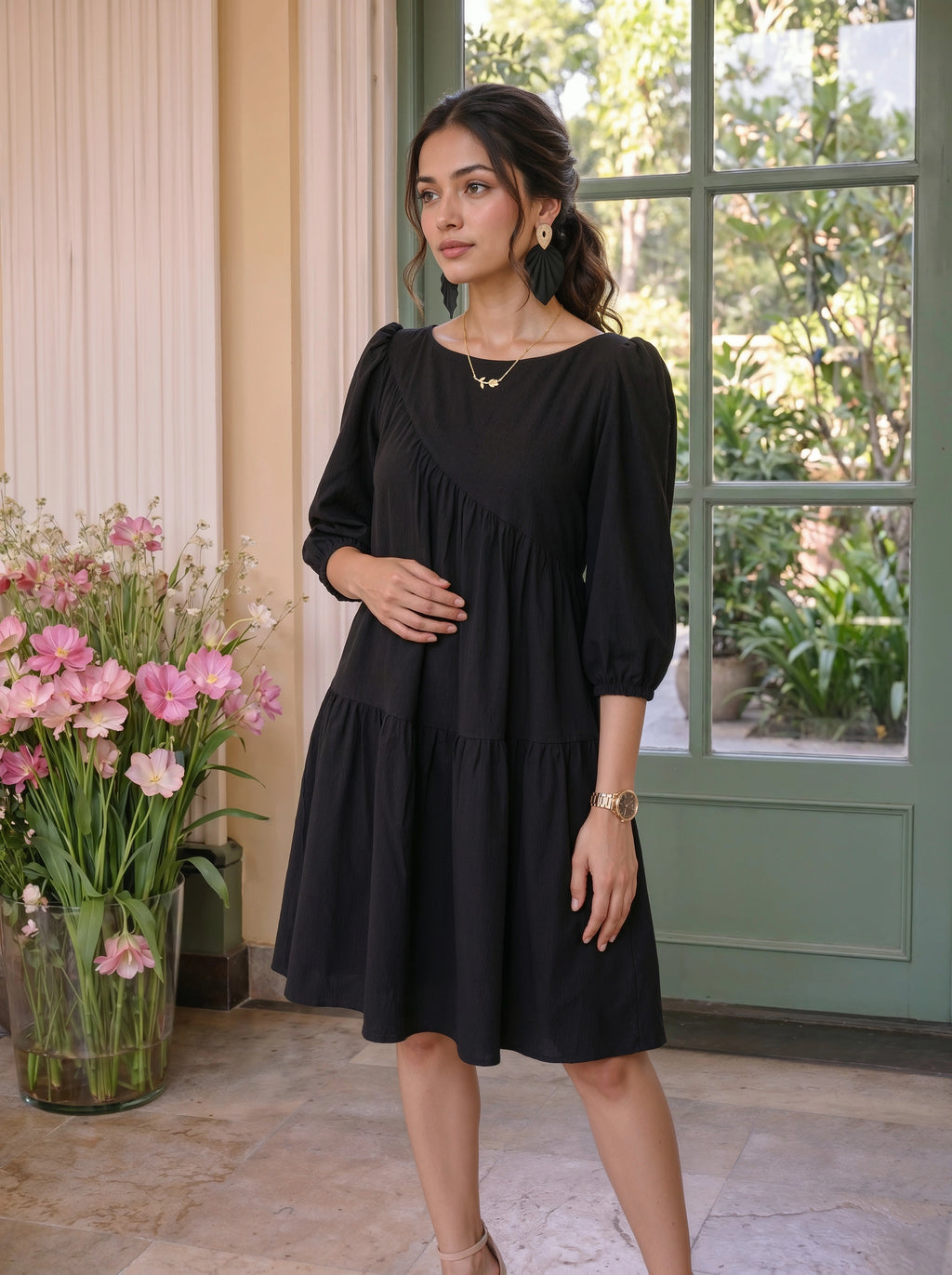Woman in a black dress standing in a room with flowers and a window.