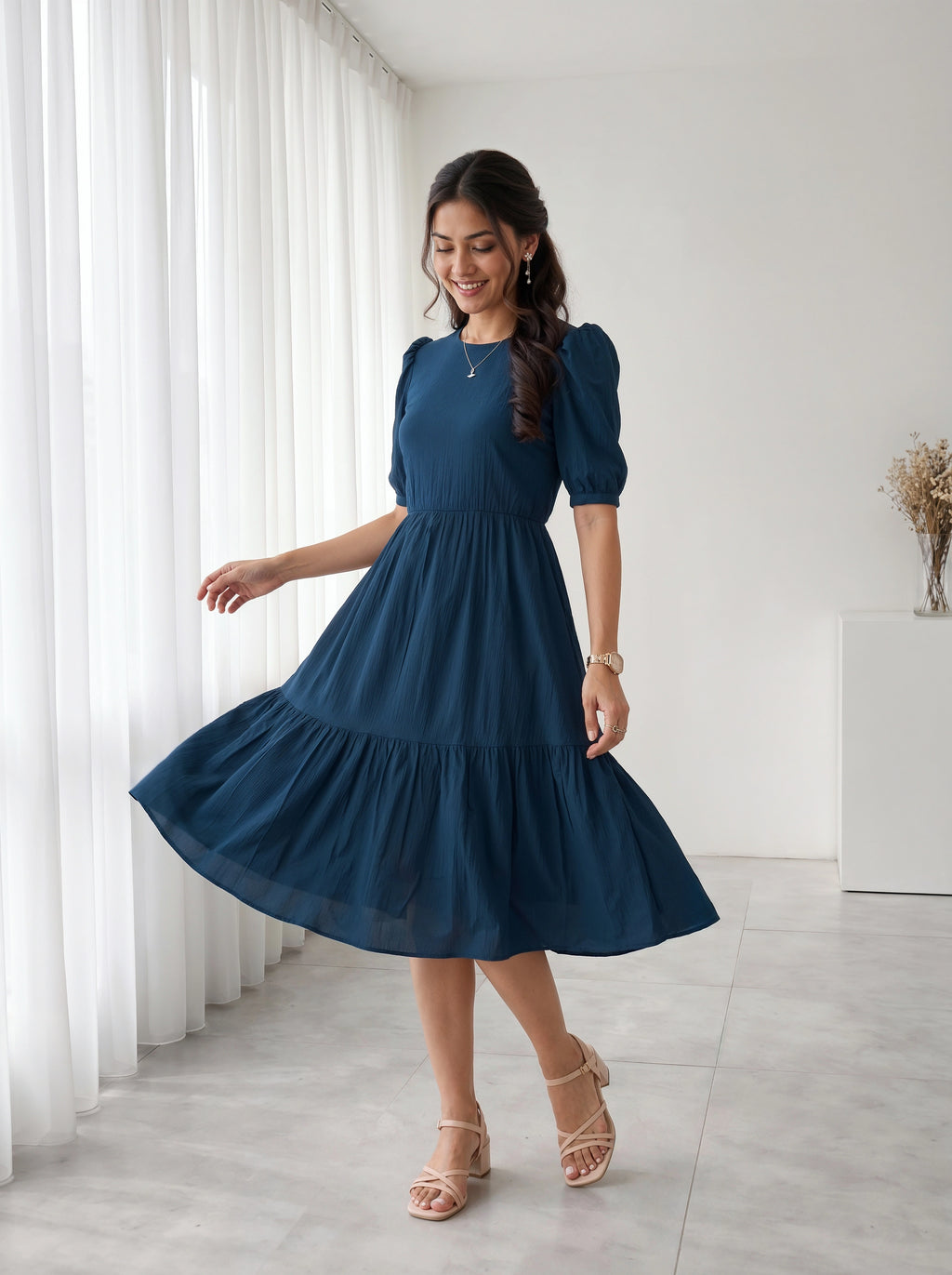 Woman in a blue dress standing in a room with white curtains and a white wall.