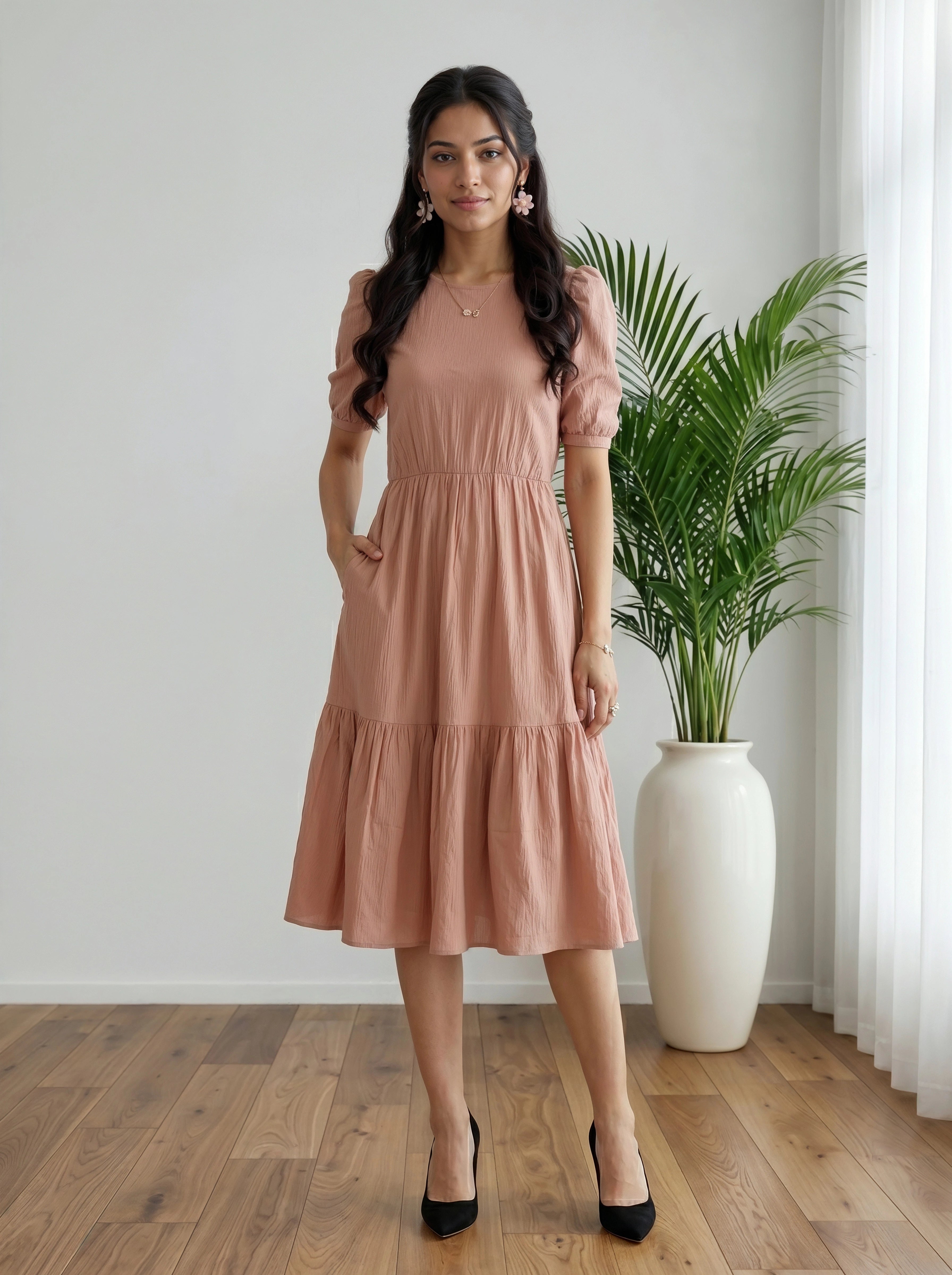 Woman wearing a pink dress standing in a room with a plant and white curtain.