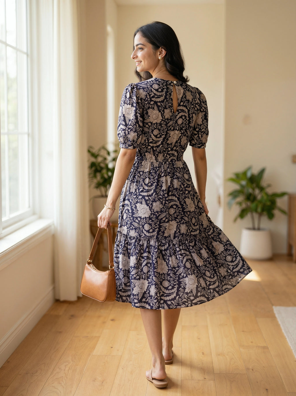 Woman in a blue floral dress holding a brown handbag in a room with wooden floors and plants.
