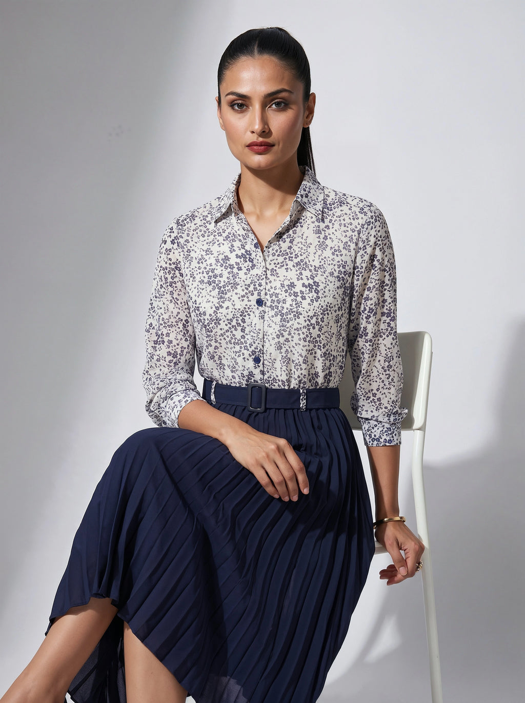 Woman wearing a floral blouse and navy skirt sitting on a chair against a gray background
