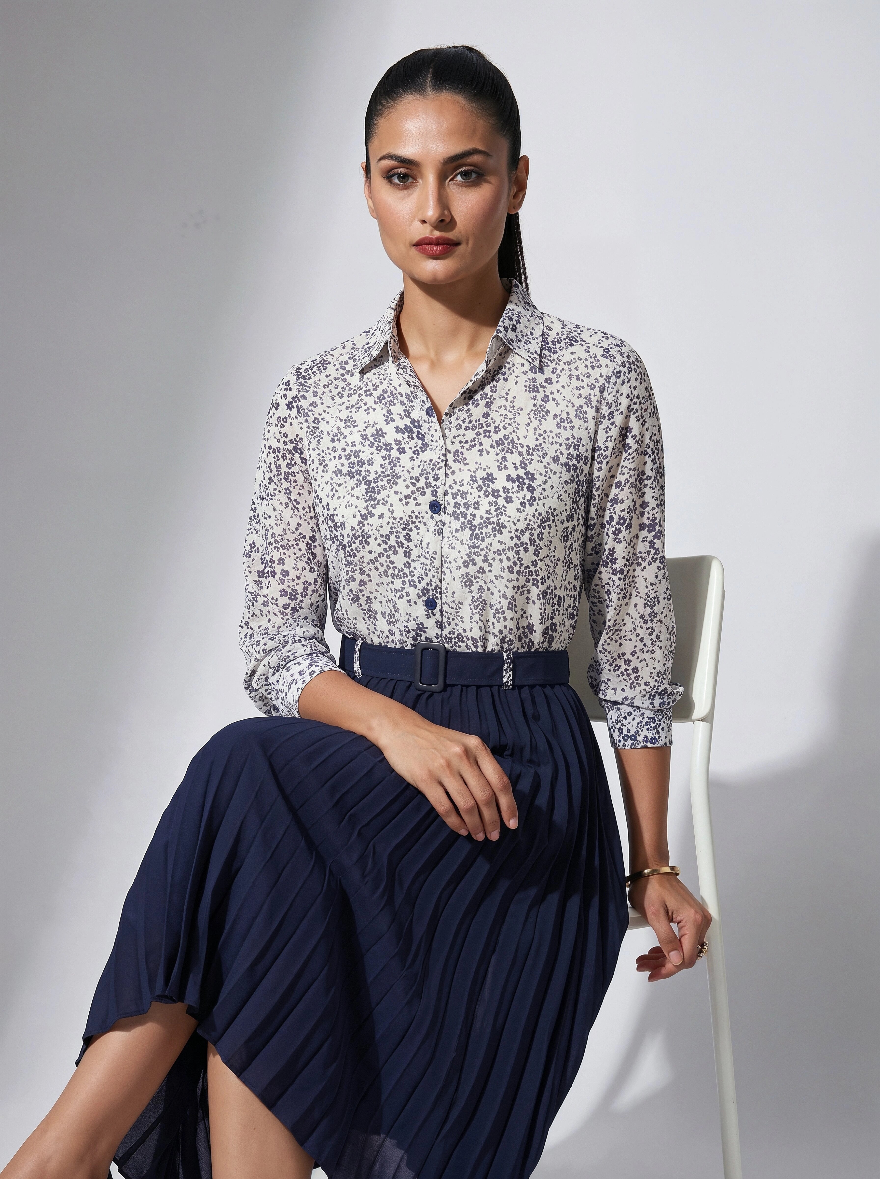 Woman wearing a floral blouse and navy skirt sitting on a chair against a gray background