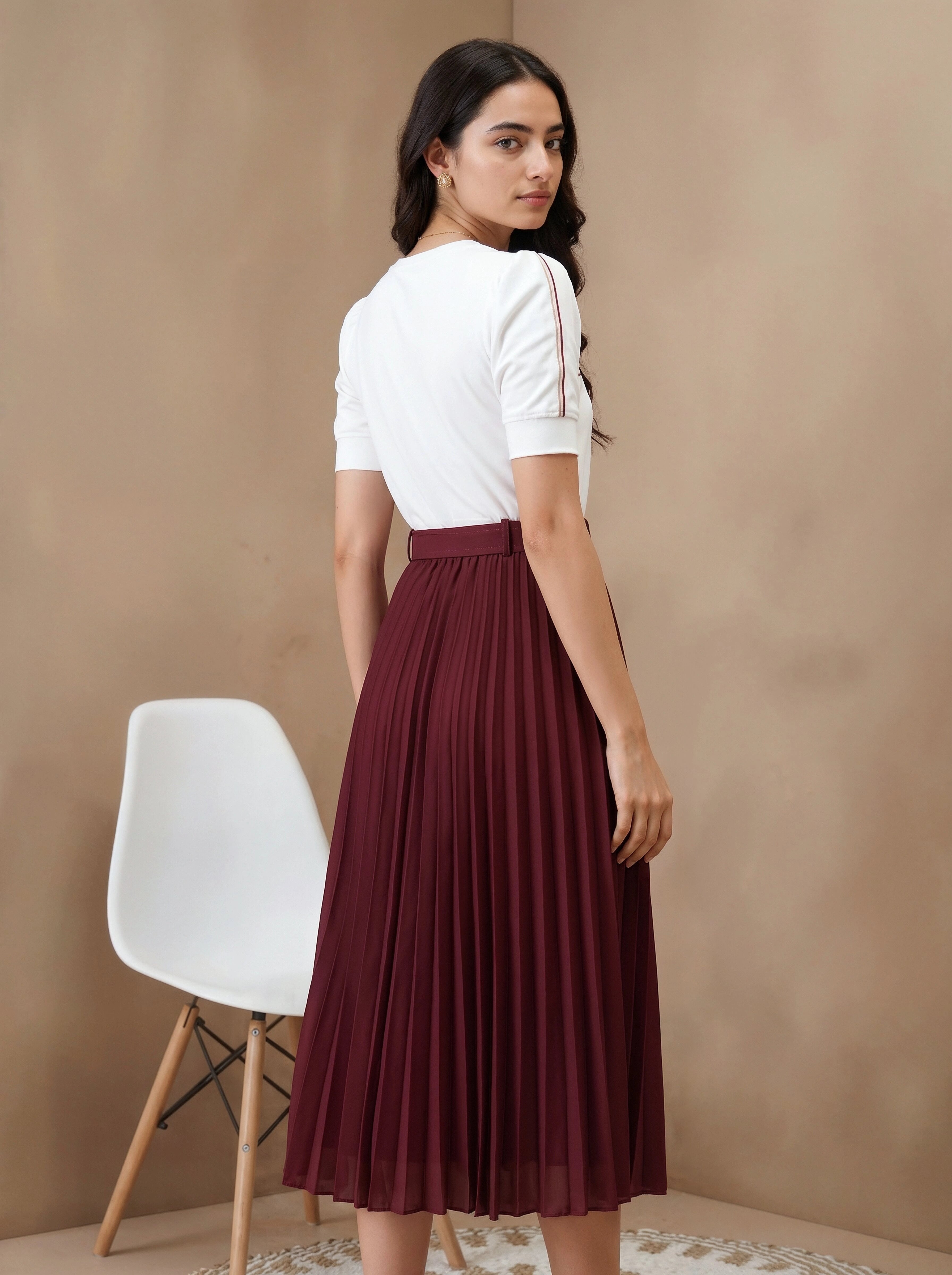 Woman wearing a white top and burgundy skirt standing against a beige wall with a white chair.