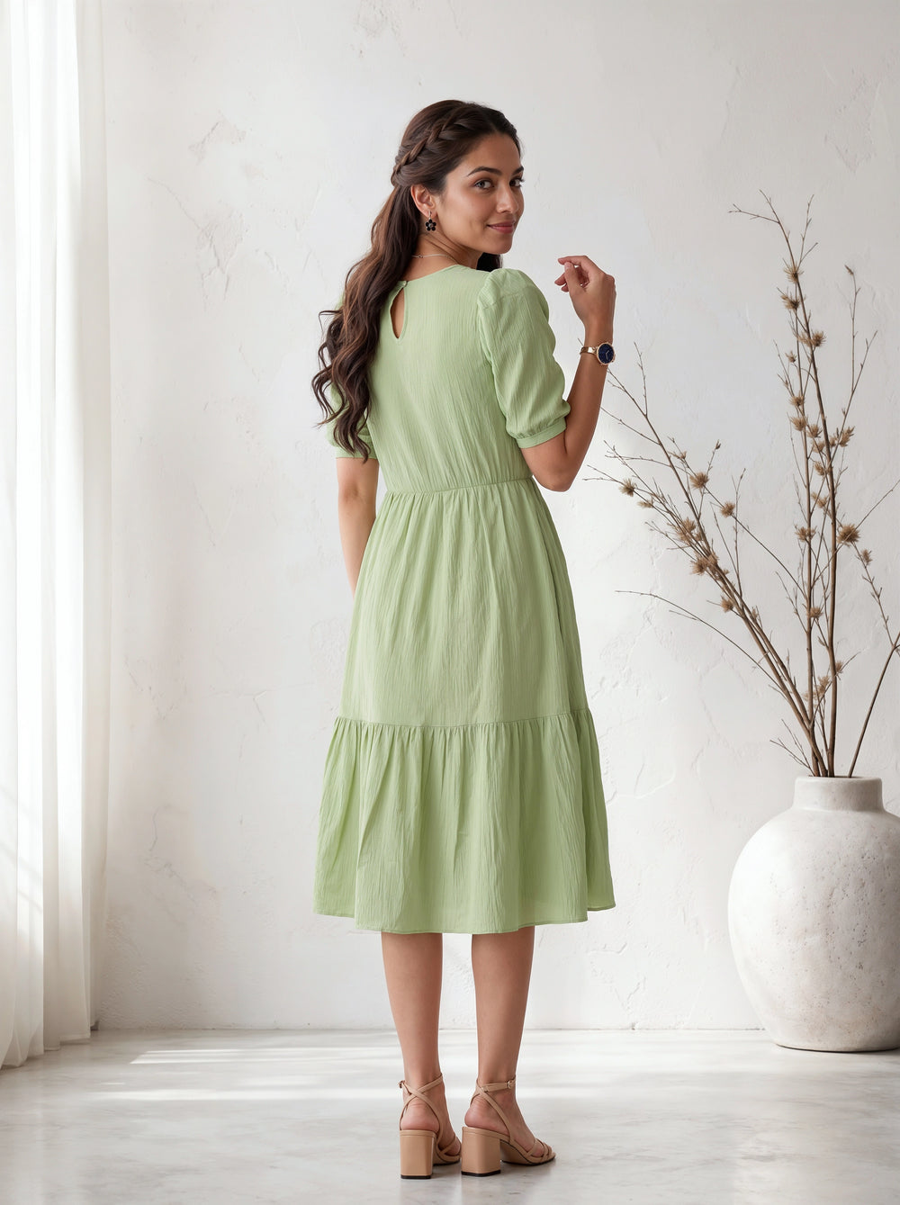 Woman wearing a light green dress standing in a bright room with a vase and plant in the background.