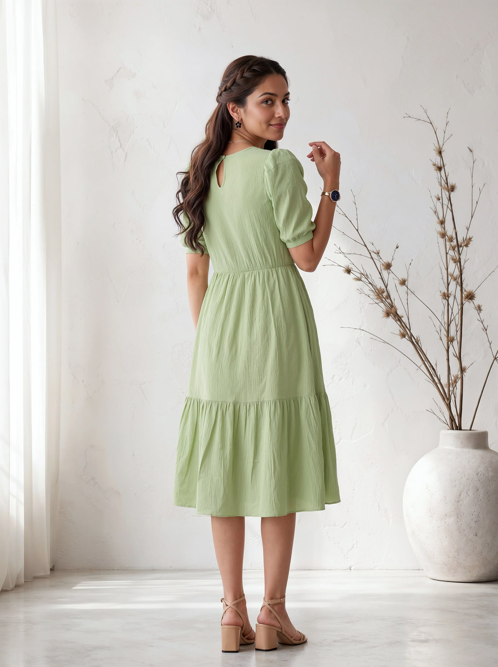 Woman wearing a light green dress standing in a bright room with a vase and plant in the background.